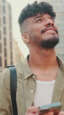 VERTICAL VIDEO: Young smiling man dressed in an olive-colored shirt stands with cellphone in his hand on modern building background