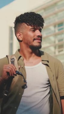 VERTICAL VIDEO: Young male student dressed in an olive colored shirt turns his head and smiles at the camera from outside next to the university