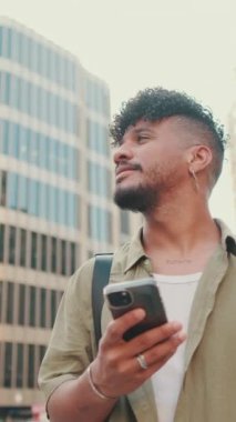 VERTICAL VIDEO: Young smiling man with beard dressed in an olive color shirt uses phone map app on the old city background