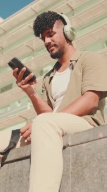 VERTICAL VIDEO: Young male student dressed in an olive color shirt sits outside next to the university, listens to music on headphones, selects tracks on his phone