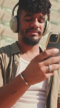 VERTICAL VIDEO: Close up, young male student dressed in an olive color shirt sits outside next to the university, listens to music on headphones, selects tracks on his phone