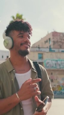 VERTICAL VIDEO: Young man with beard in an olive-colored shirt listens to music on headphones, dances