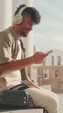 VERTICAL VIDEO: Young male student dressed in an olive color shirt sits outside next to the university, listens to music on headphones, selects tracks on his phone