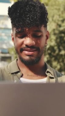 VERTICAL VIDEO: Close up, young male student wearing an olive colored shirt is working on pc laptop while sitting on bench outside the university