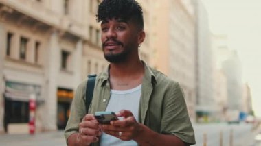 Young smiling man with beard dressed in an olive color shirt uses phone map app on the old city background