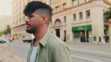Close up, young happy man with beard dressed in an olive color shirt stands next to the road, turns his head and smiles at the camera
