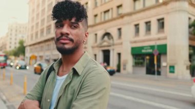 Young happy man with beard dressed in an olive-colored shirt stands next to the road, turns his head, crosses his arms and looks at the camera