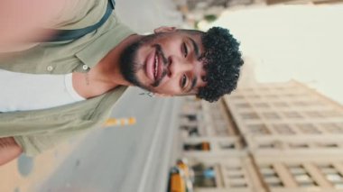 VERTICAL VIDEO: Young happy man with beard dressed in an olive color shirt stands next to the road uses smartphone, smiles and makes video call