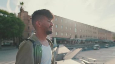 Young man dressed in an olive-colored shirt is standing in the port looking at the yachts
