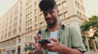 Young happy man with beard dressed in an olive-colored shirt walks through the center of the old city using mobile phone. Camera moving forwards