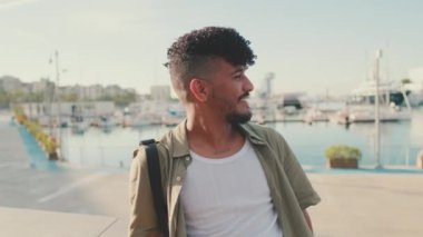 Close up, young smiling man dressed in an olive-colored shirt sits on the promenade in the port and looks at the yachts