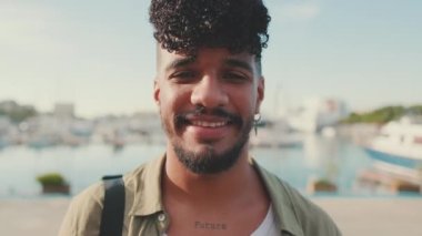 Close-up of young smiling man dressed in an olive color shirt sitting on the waterfront in the port looking at the camera