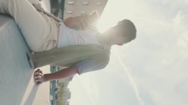 VERTICAL VIDEO: Young man dressed in an olive-colored shirt is sitting on the waterfront in the port watching videos and photos on his cellphone. Backlight