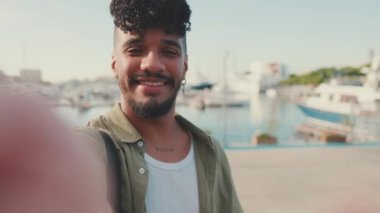 Close up, young smiling man dressed in an olive-colored shirt takes selfie while standing on the waterfront in the port