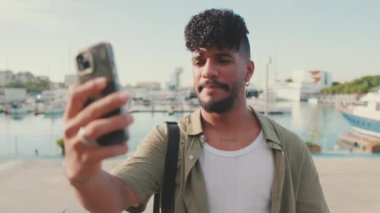 Young smiling man dressed in an olive-colored shirt takes selfie while standing on the waterfront in the port