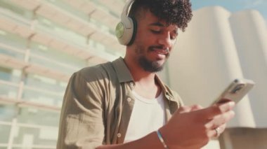 Close up, young male student dressed in an olive color shirt sits outside next to the university, listens to music on headphones, selects tracks on his phone
