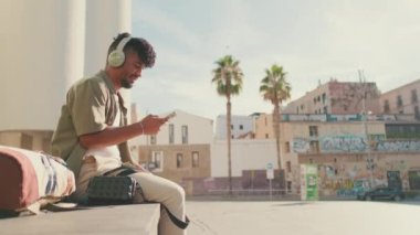 Young male student dressed in an olive color shirt sits outside next to the university, listens to music on headphones, selects tracks on his phone