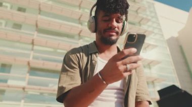 Close up, young male student dressed in an olive color shirt sits outside next to the university, listens to music on headphones, selects tracks on his phone