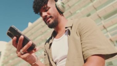 Close up, young male student dressed in an olive color shirt sits outside next to the university, listens to music on headphones, selects tracks on his phone. Camera moving forwards