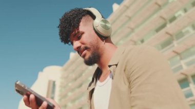 Close up, young male student dressed in an olive color shirt sits outside next to the university, listens to music on headphones, selects tracks on his phone.Camera moves backwards
