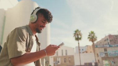 Close up, young male student dressed in an olive color shirt sits outside next to the university, listens to music on headphones, selects tracks on his phone. Camera moves backwards