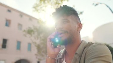 Clouse up, young man with beard wearing an olive colored shirt with headphones talking on the phone while sitting on bench on modern buildings background