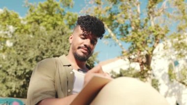 Young male student wearing an olive colored shirt takes notes in notebook while sitting on bench outside the university
