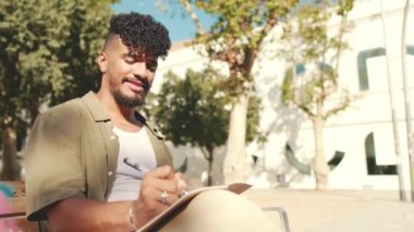 Young male student wearing an olive colored shirt takes notes in notepad while sitting on bench outside the university
