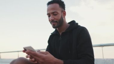 Kapüşonlu giyinmiş genç sakallı erkek sporcu antrenmandan sonra sette otururken cep telefonunu kullanıyor. Adam kafasını kaldırıp kameraya bakıyor.