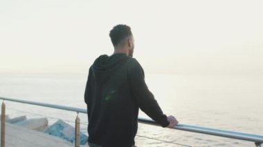 Genç sakallı erkek sporcu setin üzerinde durmuş denize bakıyor. Arka plan