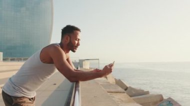 Genç sakallı erkek sporcu, sette antrenmandan sonra dinlenirken cep telefonu kullanıyor.