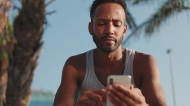 Yakından, genç sakallı erkek sporcu antrenmandan sonra dinleniyor, bankta oturuyor, akıllı telefon kullanıyor.