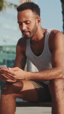 VERTICAL VIDEO: Young bearded male athlete is resting after training, sitting on bench, using smartphone on modern building background