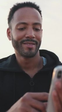 Kapüşonlu kapüşonlu sakallı, genç erkek sporcu sette otururken cep telefonunu kullanıyor.