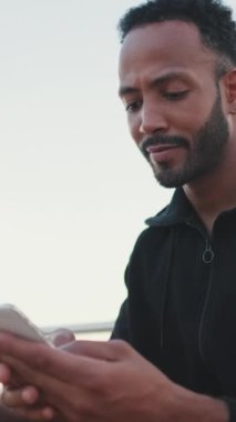 Kapüşonlu kapüşonlu sakallı, genç erkek sporcu sette otururken cep telefonunu kullanıyor.