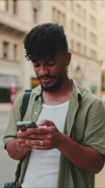 VERTICAL VIDEO: Young smiling man with beard dressed in an olive-colored shirt uses phone on an old city background. Guy reads the good news