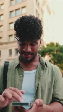 VERTICAL VIDEO: Young smiling man with beard dressed in an olive color shirt uses phone map app on the old city background