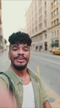 VERTICAL VIDEO: Young happy man with beard dressed in an olive color shirt stands next to the road uses smartphone, smiles and makes video call