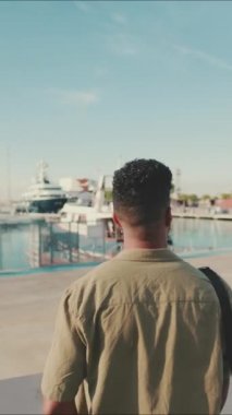 VERTICAL VIDEO: Young man dressed in an olive-colored shirt is standing in the port looking at the yachts. Back view