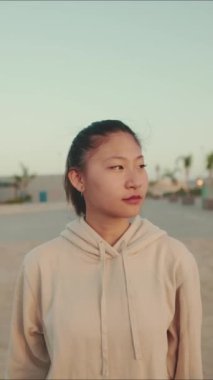 VERTICAL VIDEO: Asian girl in sportswear uses the phone while standing on the waterfront at morning time