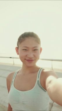 VERTICAL VIDEO: Smiling asian girl in sports top makes selfie while standing on the embankment in the morning light