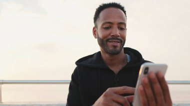 Yaklaş, kapüşonlu giyinmiş genç sakallı erkek sporcu antrenmandan sonra sette otururken cep telefonunu kullanıyor.