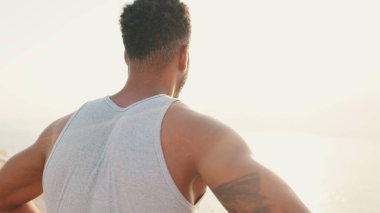 Genç sakallı, formda bir sporcu, setin üzerinde durmuş denize bakıyor..