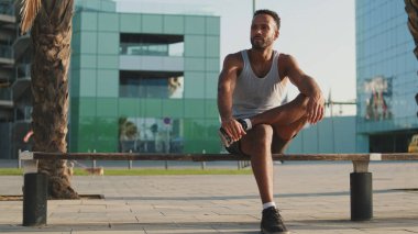 Genç sakallı erkek sporcu, eğitimden sonra modern arka planda bankta dinleniyor.