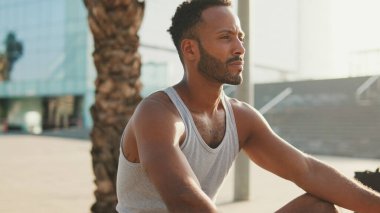 Genç sakallı erkek sporcu, eğitimden sonra modern arka planda bankta dinleniyor.