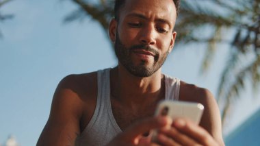 Genç sakallı erkek sporcu antrenmandan sonra dinleniyor, bankta oturuyor, akıllı telefon kullanıyor. 