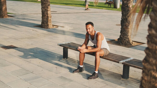 Young bearded male athlete is resting after training, sitting on bench, using smartphone 