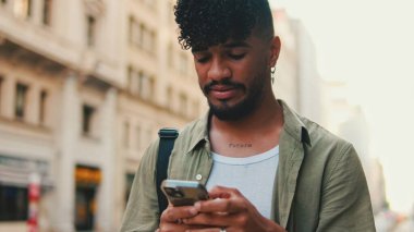 Young smiling man with beard dressed in an olive color shirt uses phone map app on the old city background