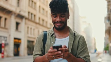 Young smiling man with beard dressed in an olive color shirt uses phone map app on the old city background