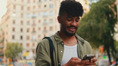 Young smiling man with beard dressed in an olive color shirt uses phone map app on the old city background